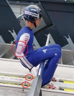 Skispringer mit blauem Anzug und Helm beim Start auf der Piste; der Springer trägt Inertialsensoren und Kraftmesssohlen, die im Bild rot markiert sind 