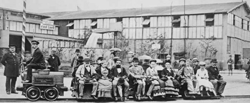 Schwarz-weiß Aufnahme der ersten elektrischen Lokomotive und vielen Personen, im Hintergrund ist ein Gebäude zu sehen